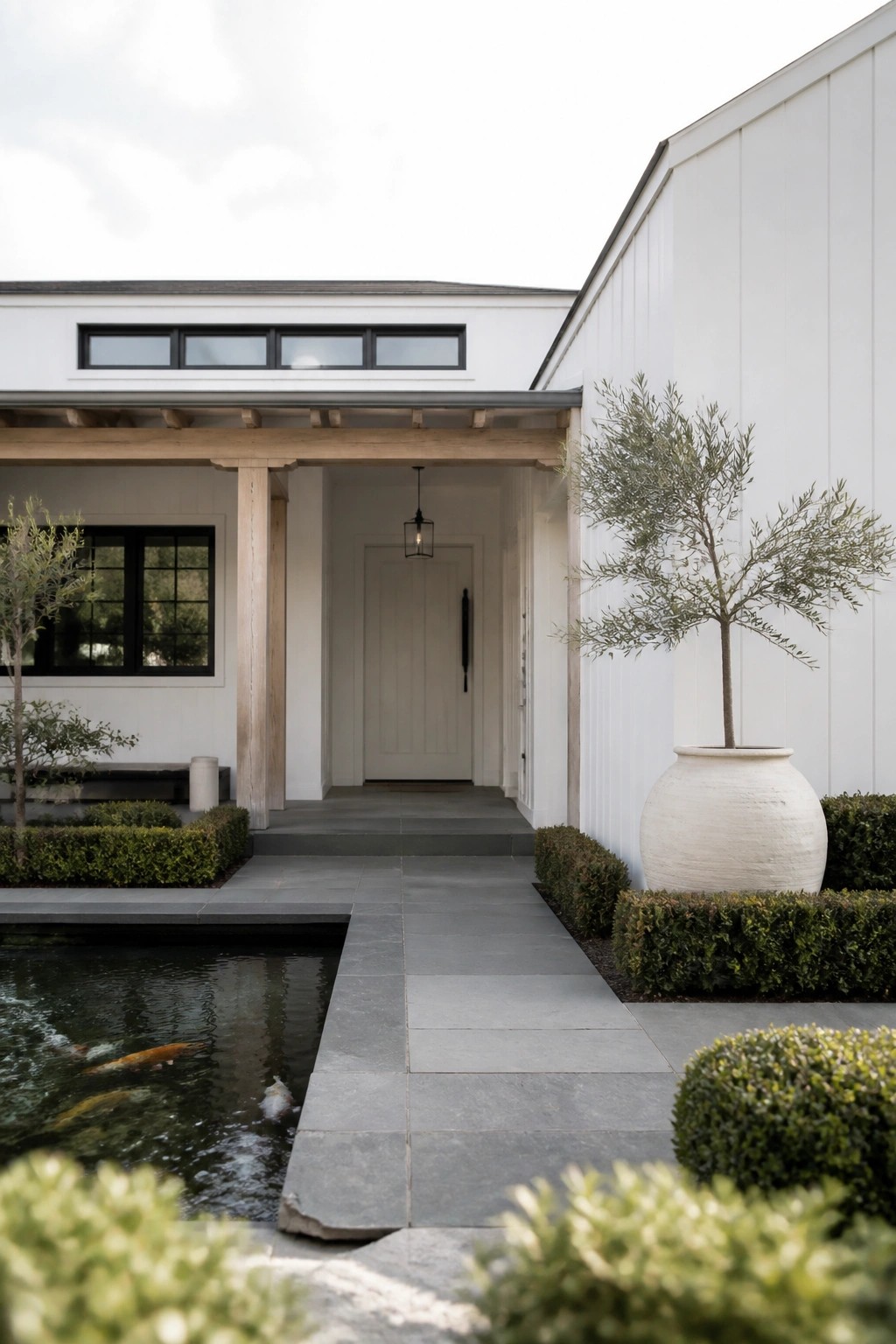 A contemporary white house with black-framed windows, a wooden entry overhang, a stone pathway, and a koi pond beside neatly trimmed shrubs.
