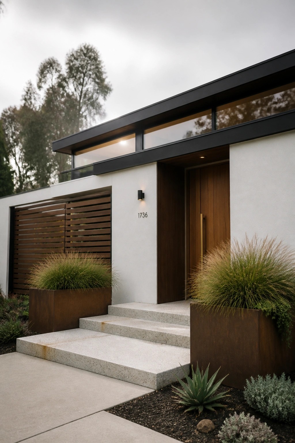 Modern house entrance with white walls, dark trim, wood door, horizontal clerestory windows above, concrete steps, and metal planters with grasses.
