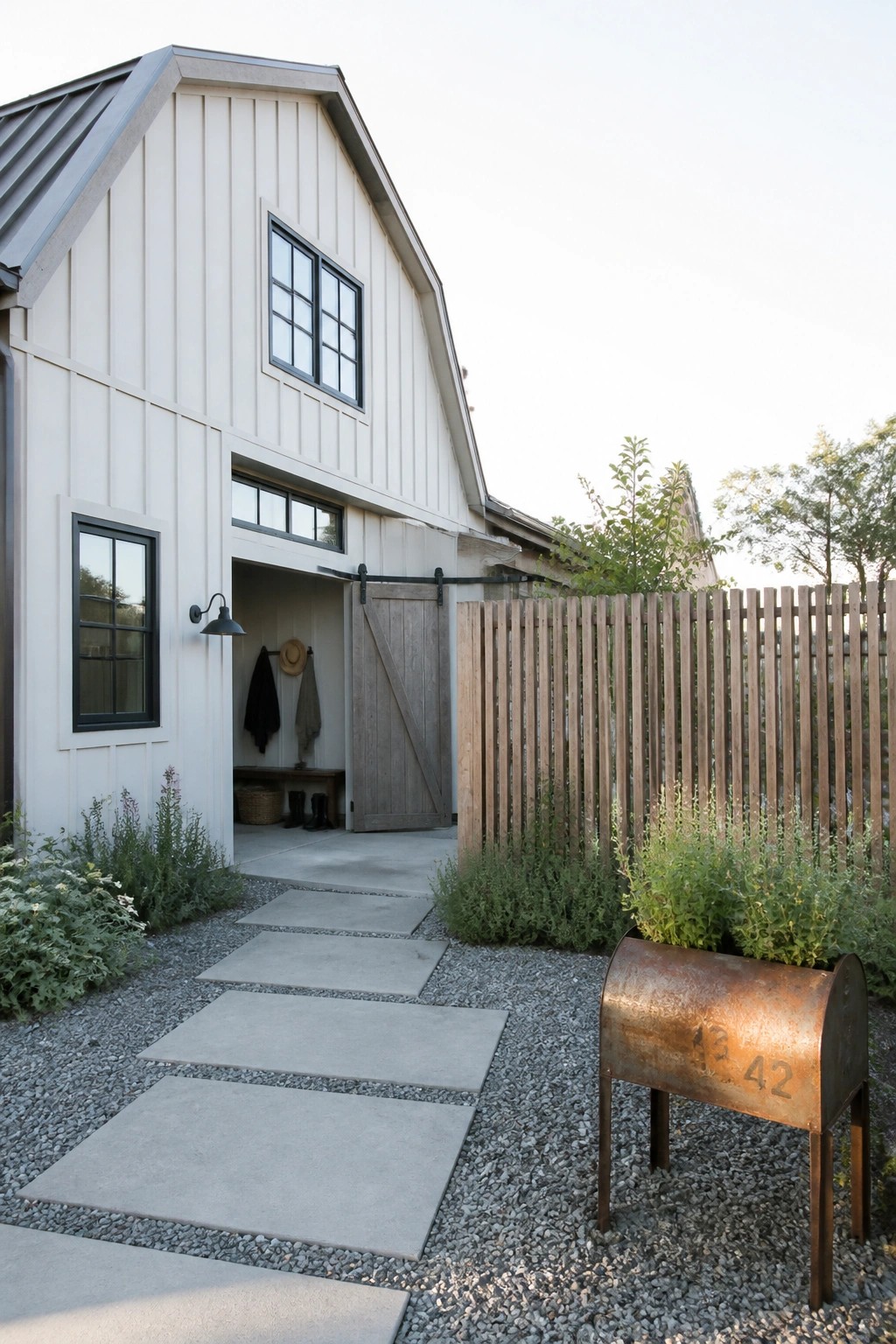 A modern farmhouse exterior featuring white vertical siding, a large sliding wooden barn door at the entrance with black hardware, black-framed windows, a gravel path with concrete pavers, and a rusted metal mailbox in the foreground.