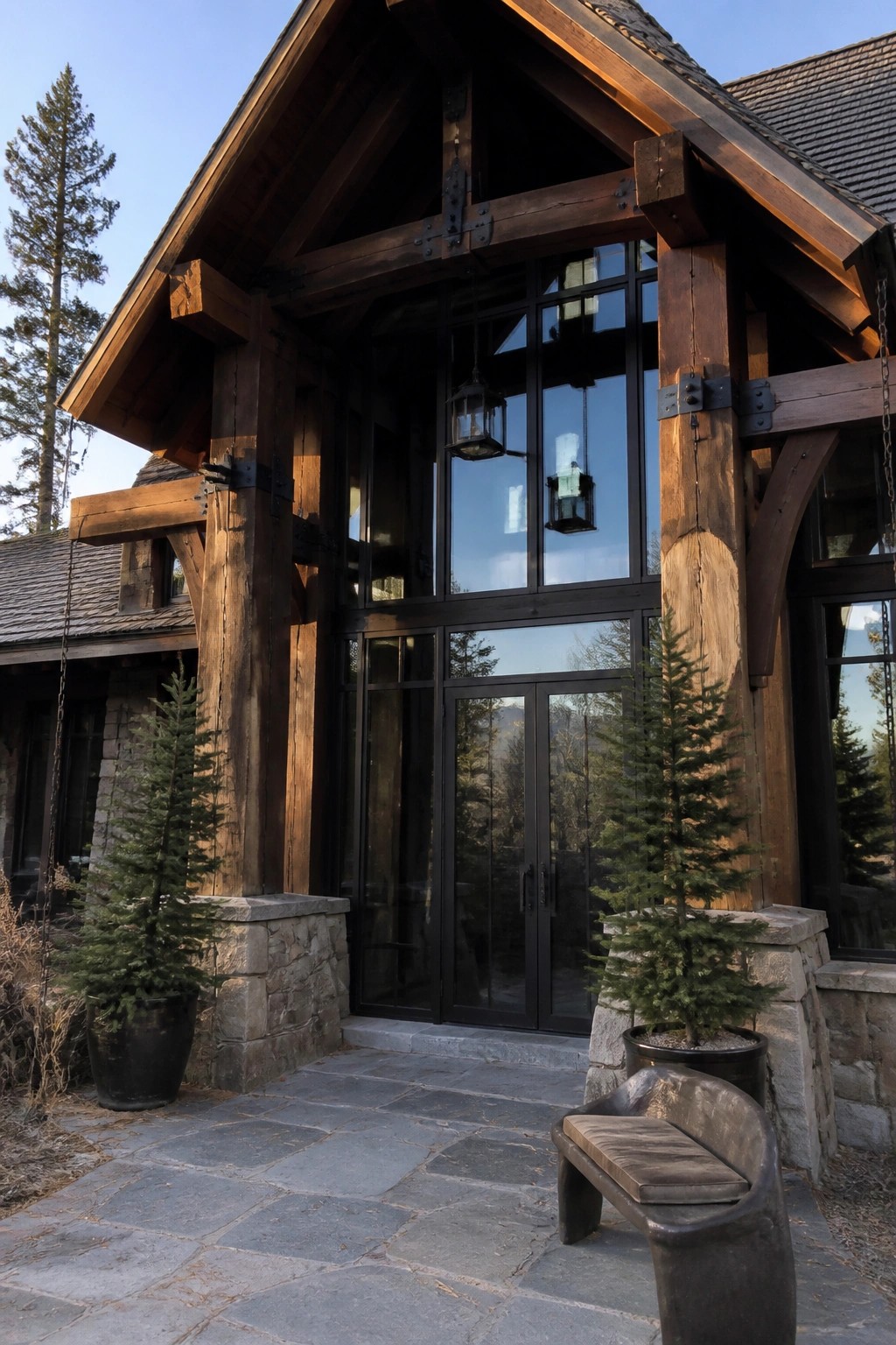 A rustic wooden house entrance features tall black-framed glass doors and windows, flanked by potted evergreen trees on stone bases and a curved bench on a stone patio.