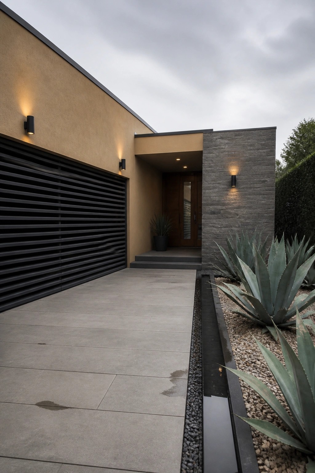 Modern house exterior with a large black horizontal-slatted garage door to the left of a recessed entry featuring a wooden door, stone accent wall, and concrete walkway with agave plants.