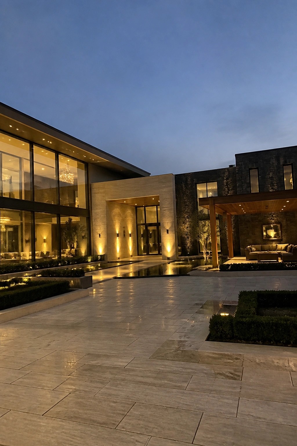 Modern luxury home with illuminated glass windows, stone courtyard, and reflecting pool at dusk.