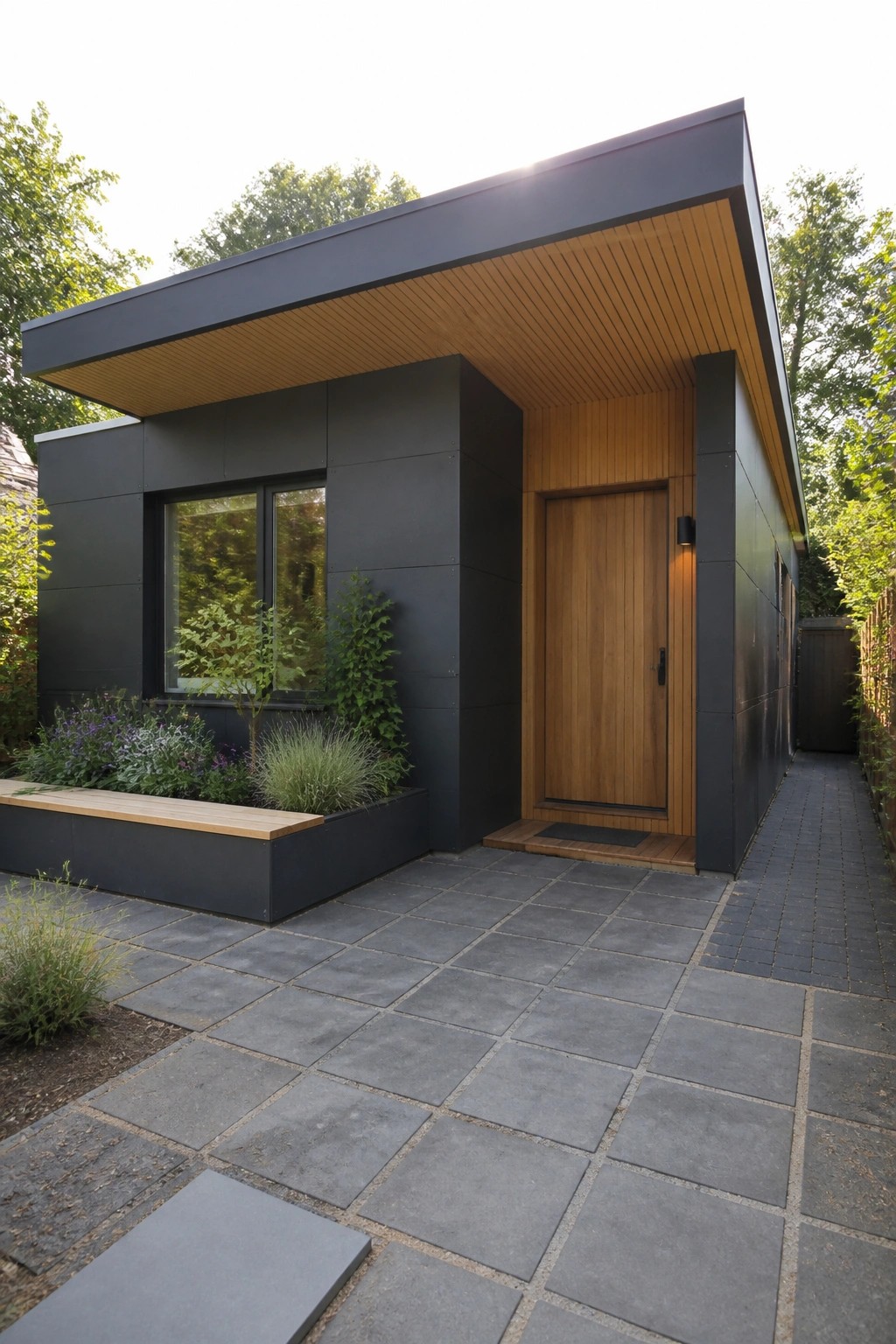 A modern house with dark gray siding, a deep roof overhang with wood underside, a wooden front door, and a paved stone entry area with nearby plants.