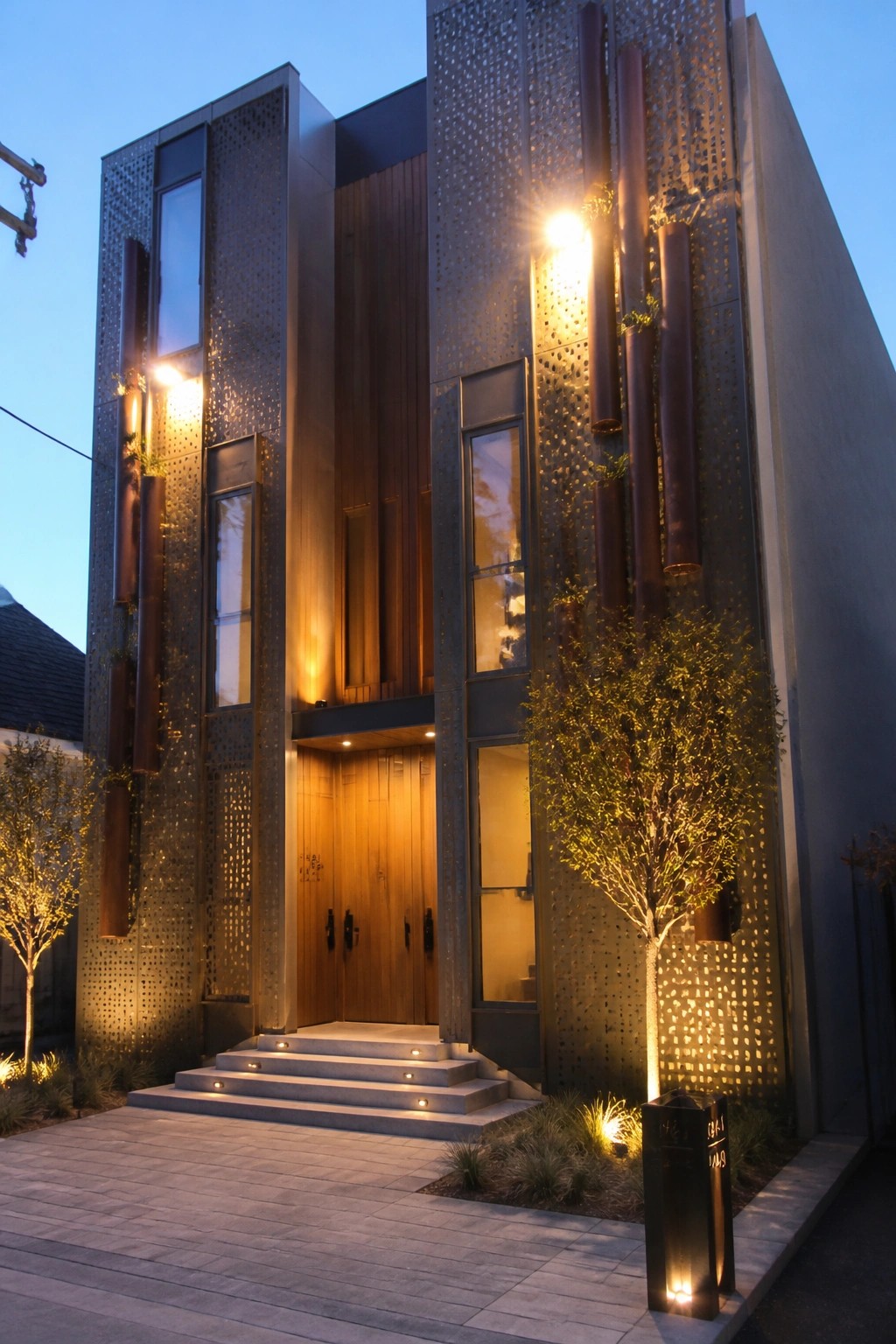 A modern two-story house at dusk with tall perforated metal panels, vertical light columns, wooden entry doors, and concrete steps.