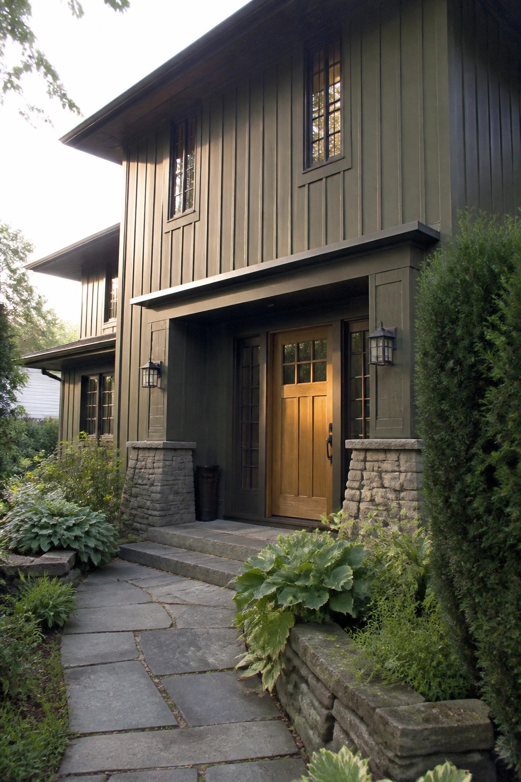 A two-story house with dark green vertical siding, a wooden front door under a covered entry, stone pillars, and a stone pathway leading to the steps.