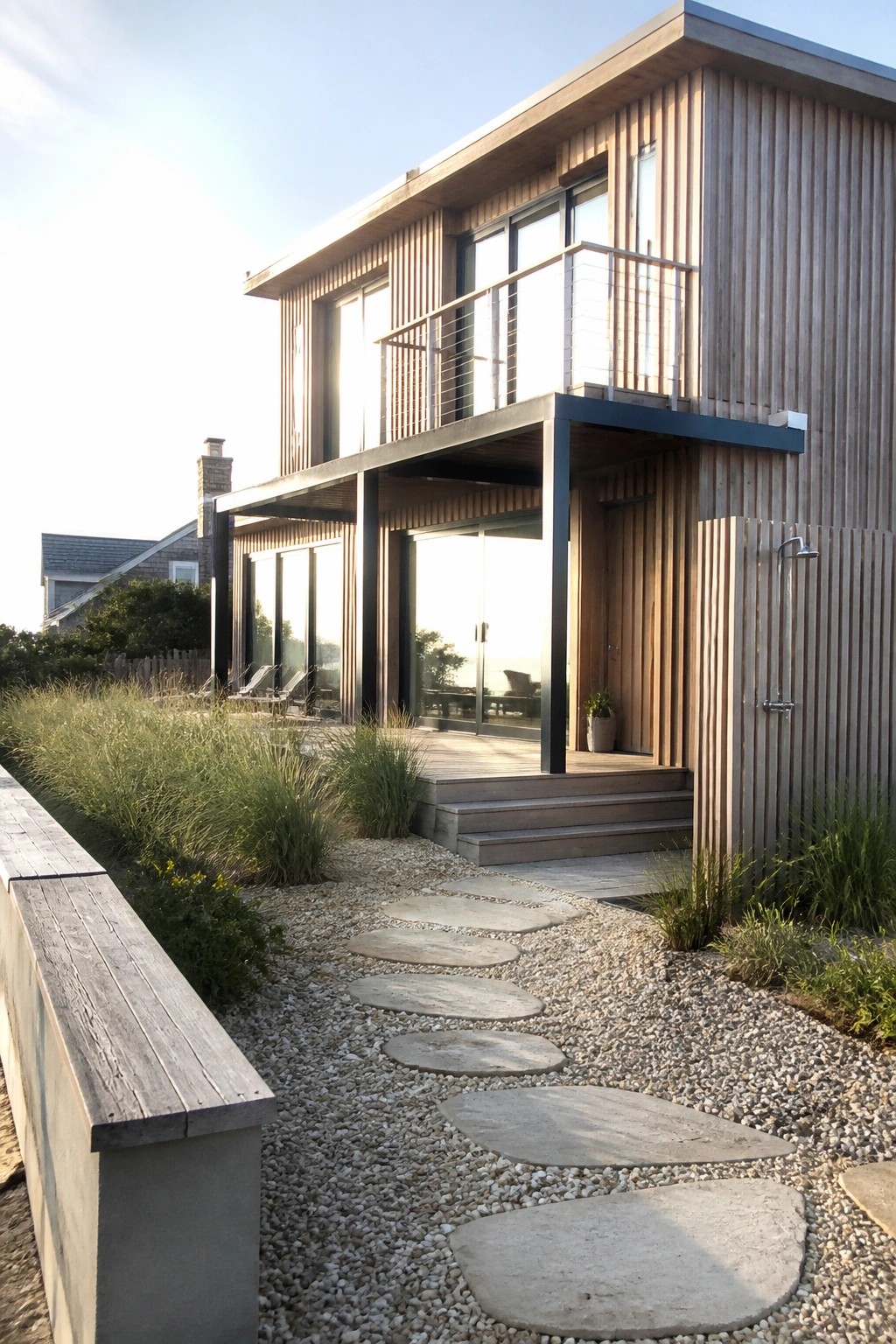 A modern two-story house clad in vertical wood siding with a cantilevered upper level, balcony, and stepping stone path through gravel.