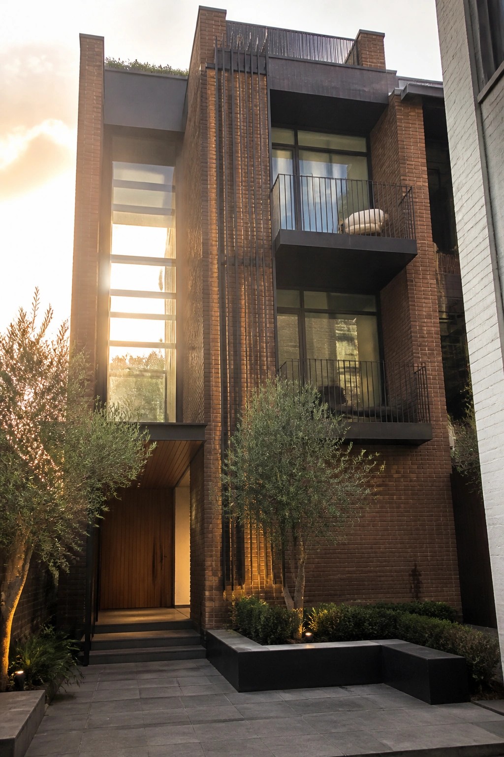 Modern brick townhouse with balconies, large windows, wooden door, and trees at sunset.